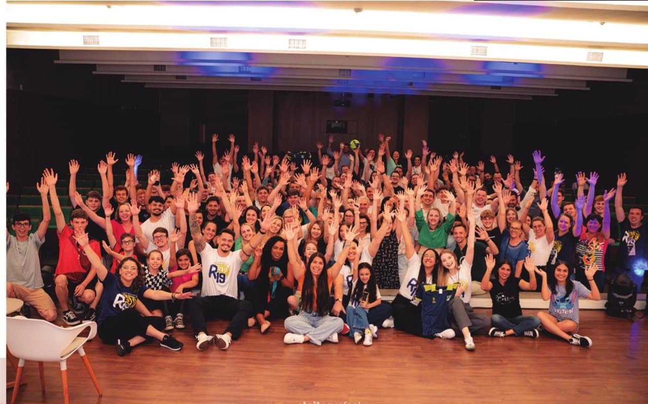 Foto de grupo no auditório, com dezenas de alunos, famílias e professores de braços erguidos