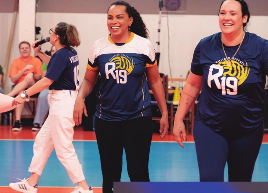 Três professoras com a camisa do R19 caminham sorrindo pela quadra