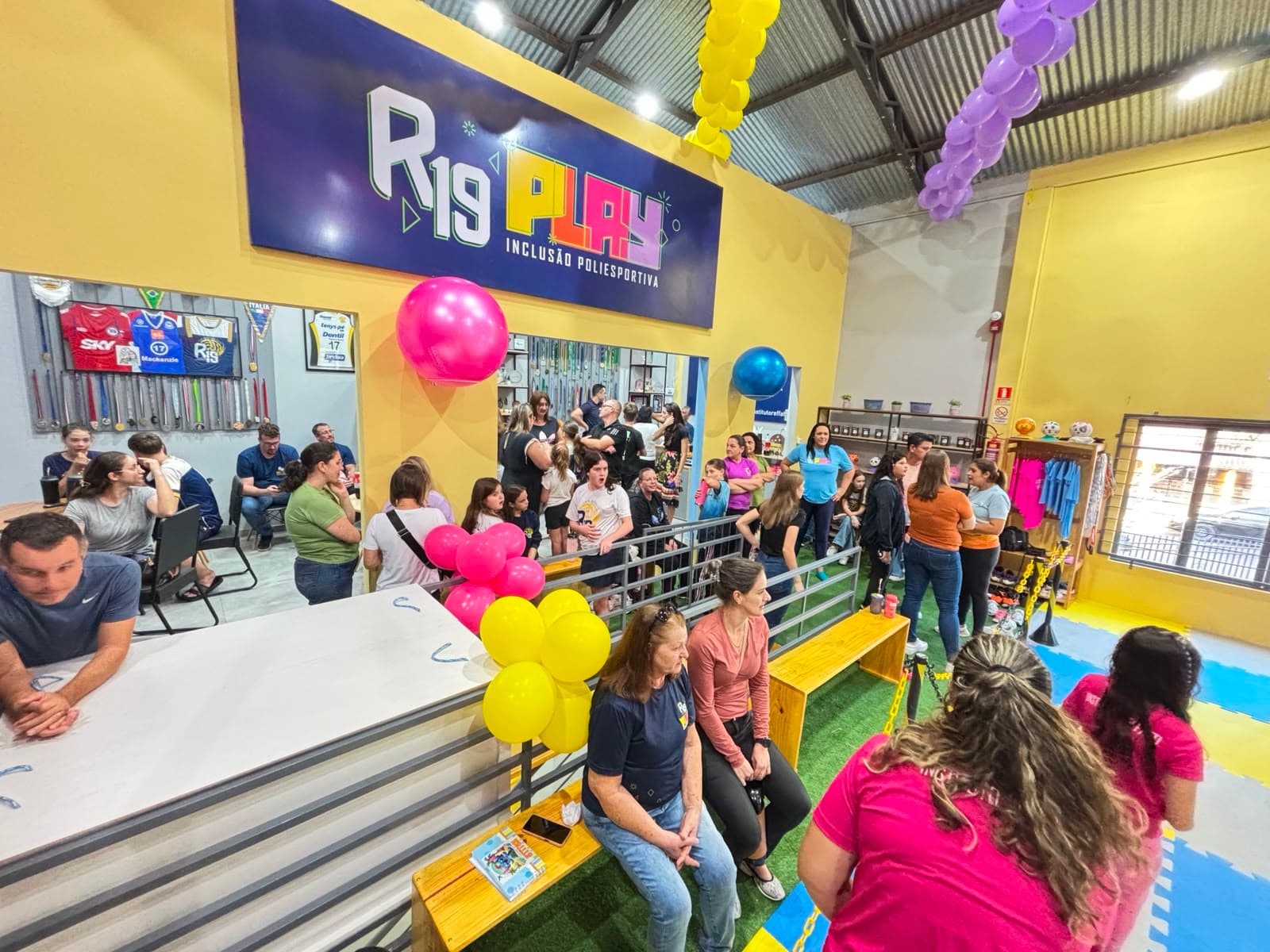 Salão do R19 PLAY sob a placa 'R19 PLAY — Inclusão Poliesportiva', com balões coloridos, piso de grama sintética e famílias reunidas no espaço