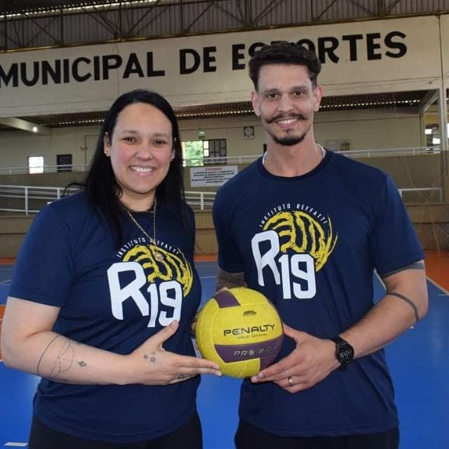 Willian Reffatti e Ju Costa lado a lado no Ginásio Municipal de Esportes, de camiseta do R19 e segurando juntos uma bola de vôlei